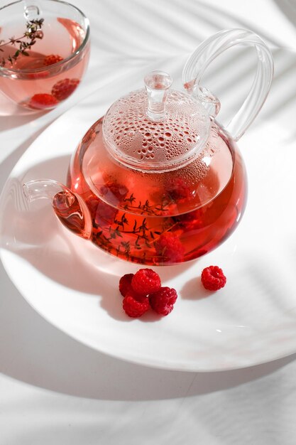 Roter Beerenkräutertee mit Thymian und frischer Himbeere in Teekanne aus Glas. Weißer Hintergrund mit Exemplar