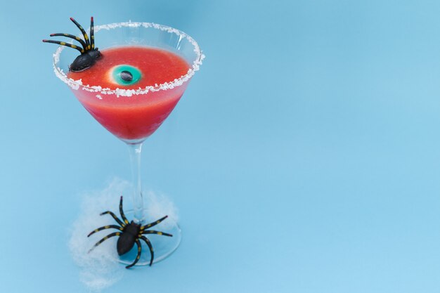 Roter Cocktail im Glas verziert für Halloween mit Spinne und Auge auf blauem Hintergrund. Platz kopieren.