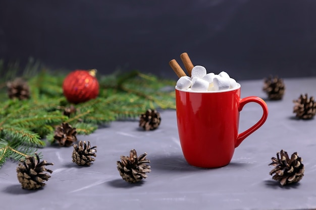 Roter Kakaobecher mit Marshmallows und Zimtstangen, Fichtenzweigen, Zapfen und einer Weihnachtskugel