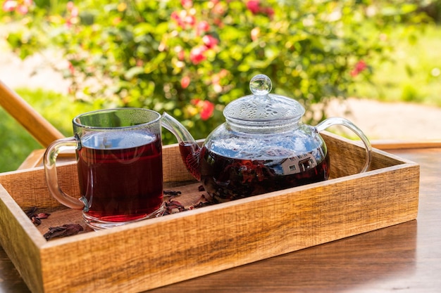 Roter Karkade-Tee in einer Teekanne aus Glas und Tasse auf einer Holzkiste im Garten