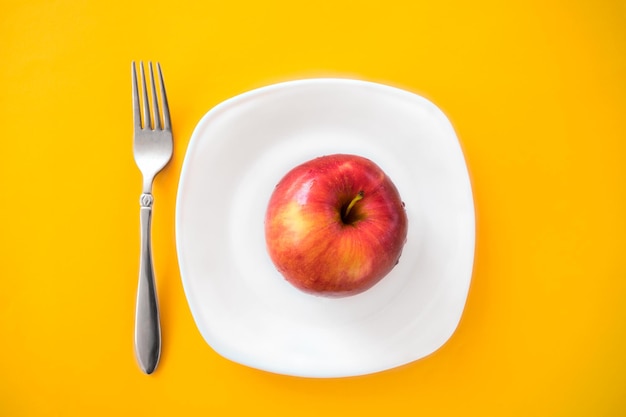 Roter reifer Apfel liegt in einem weißen Teller mit einer Gabel auf gelbem Hintergrund Richtige Ernährung Diät die richtige Lebensweise minimalistisches Foto über Lebensmittel