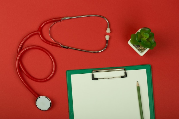 Roter Stethoskopstift am Arbeitsplatz des Arztes und leeres grünes Klemmbrett auf rotem Papierhintergrund