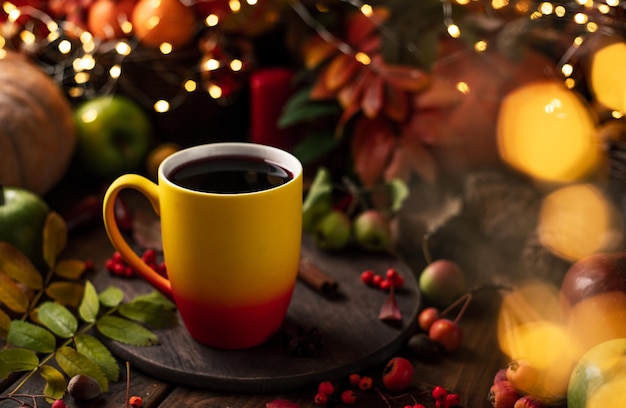 Roter und gelber Becher mit Glühwein. Früchte und Gewürze liegen überall auf einem Holztisch. Dahinter brennen gelbe Girlandenlichter