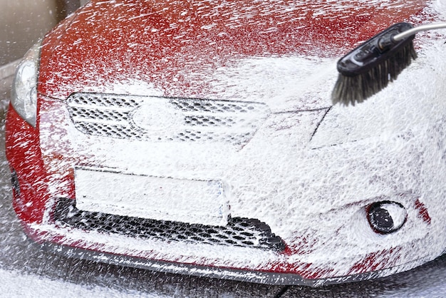 Rotes Auto in Selbstbedienungswaschanlage gewaschen, Detail auf weißem Seifenschaum, der aus Hochdruckschlauch sprüht und die gesamte vordere Stoßstange und das Nummernschild bedeckt