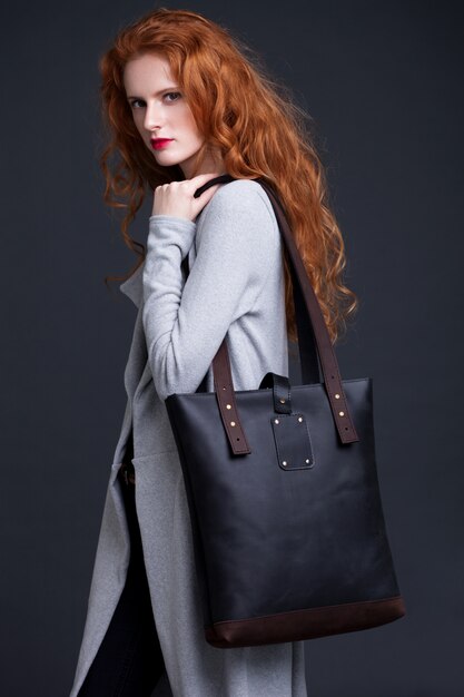 Rotes Haarmode-modell, das große dunkelblaue Ledertasche auf dunklem Hintergrund hält. Mädchen, das langen blauen Pullover trägt.