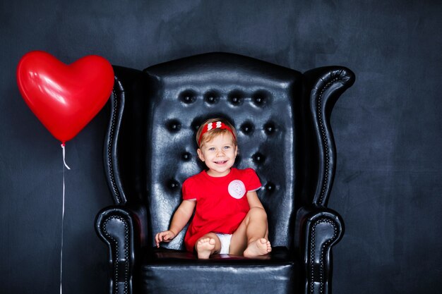 Rotes Kleid des kleinen blonden Girlins mit rotem Kranz mit den Herzen, die auf dem Lehnsessel mit rotem Herzballon am Valentinstag sitzen