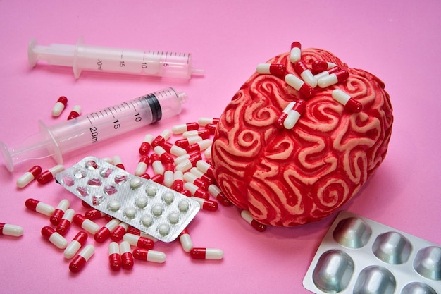 Rotes menschliches Gehirn mit zwei Spritzen und einem Haufen roter und weißer Pillen und Kapseln auf einem rosa Tisch