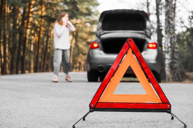 Rotes Notdreieck mit verschwommenem Auto und Frau, die Automechaniker im Hintergrund anruft, kaputtes Auto