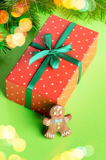 Rotes Rechteck in Tupfen verpackte Box mit Lebkuchenmann auf grünem Hintergrund mit Weihnachtsbeleuchtung. Geschenk unter Weihnachtsbaum. Weihnachten festliche Dekoration. Nahansicht. Vertikale Ausrichtung.