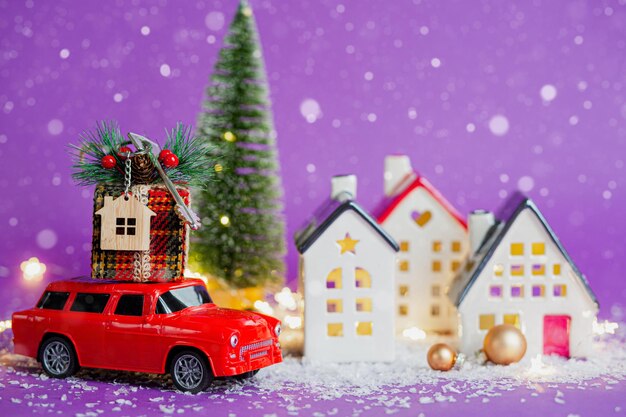 Rotes Retro-Auto auf Schnee trägt Geschenkbox mit Hauptschlüssel mit Schlüsselbundhütte auf dem Dach vorbei an Häusern mit Lichterketten, Weihnachtsbaum. Violetter Hintergrund. Gemütliches neues Jahr. Immobilien, Umzug, Hypothek.