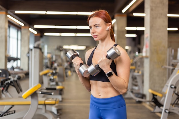 Rothaarige, fitte Frau, die in einem modernen Fitnessstudio mit Hanteln in den Händen trainiert