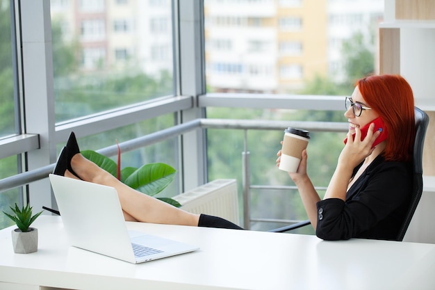 Rothaarige Frau, die am Handy spricht und remote am Laptop arbeitet