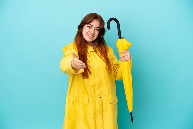 Rothaarige Frau, die einen Regenschirm isoliert auf blauem Hintergrund hält, der Hände schüttelt, um ein gutes Geschäft abzuschließen