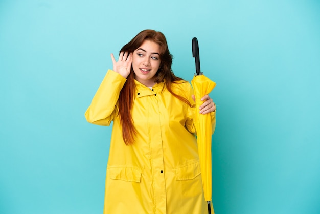 Rothaarige Frau, die einen Regenschirm isoliert auf blauem Hintergrund hält und etwas hört, indem sie die Hand auf das Ohr legt