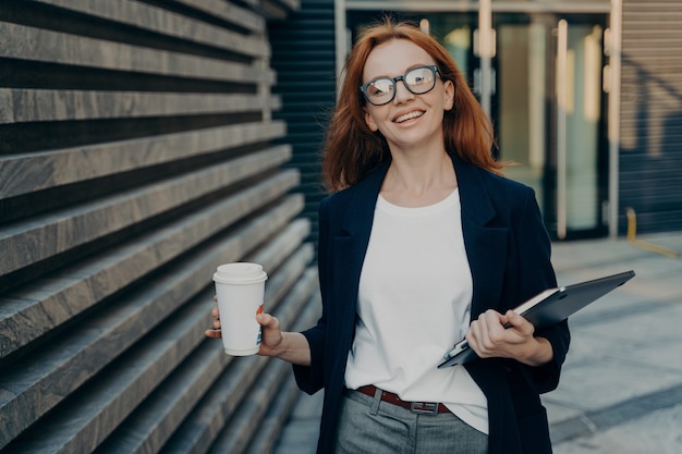 Rothaarige Frau genießt einen Spaziergang im Freien Getränke zum Mitnehmen Kaffee trägt ein digitales elektronisches Gerät Notizblock