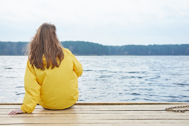 Rothaarige Frau im gelben Regenmantel sitzt auf dem Pier des Sees und schaut weit weg.