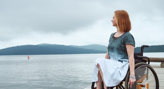 Rothaarige Frau im Rollstuhl, die schönen natürlichen Meerblick genießt. Bild mit Kopienraum für Ihre Anzeige. Banner mit textfreiraum