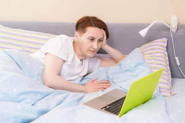 Rothaarige Frau im weißen Schlafanzug liegt im Bett unter der Decke und sieht sich einen Film auf einem Laptop an