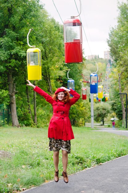 Rothaarige Frau in einem roten Barett, Mantel, der im Herbstpark spaziert, lächelt, das Leben genießt. Seilbahn. Herbst