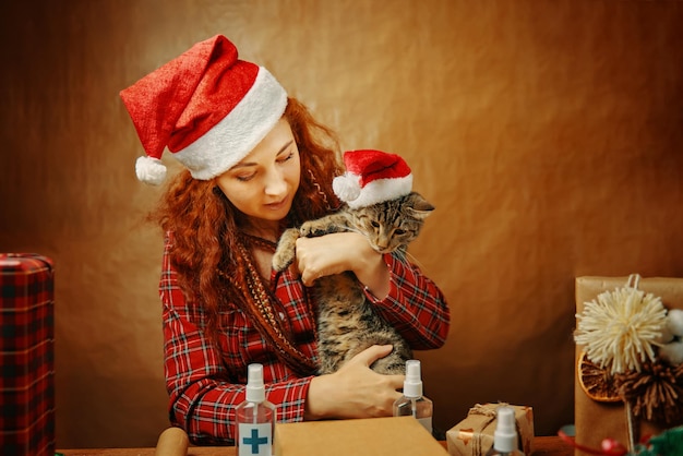 Rothaarige Frau in Weihnachtsmütze mit Katze in Weihnachtsmann-Mütze am Tisch mit Geschenkboxen und Antisept...