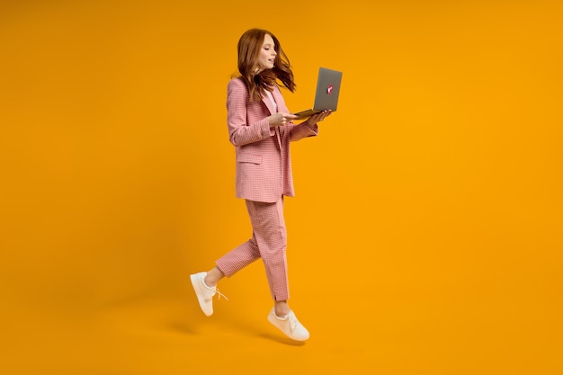 Rothaarige Frau läuft Sprung tippen Laptop trägt eleganten rosa Anzug isoliert auf gelbem Hintergrund im Studio, beeilen Sie sich. Seitenansicht Porträt der Dame am Laptop arbeiten. Kopieren Sie Platz für Werbung.