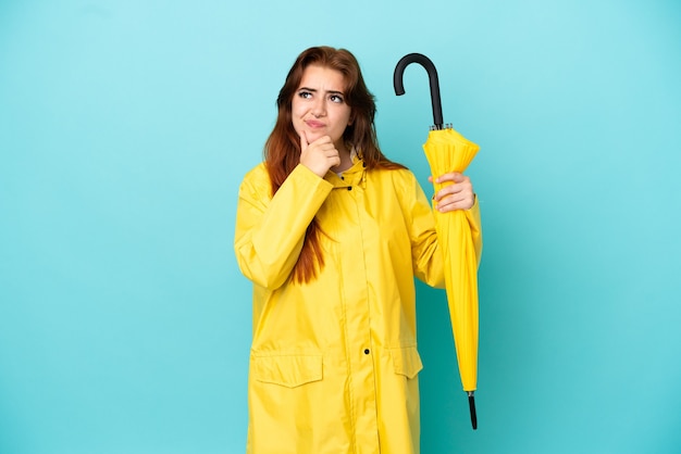 Rothaarige Frau mit einem Regenschirm isoliert auf blauem Hintergrund mit Zweifeln