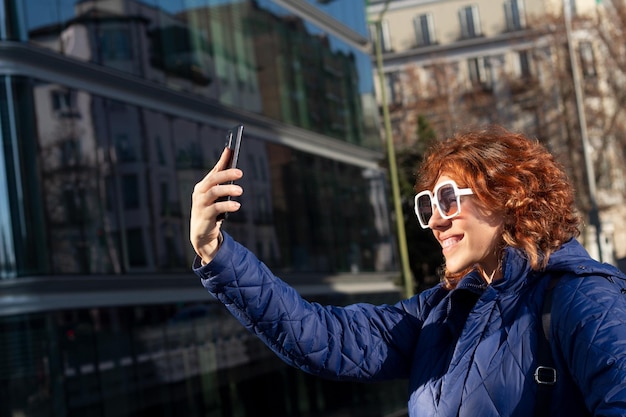 Rothaarige Frau mit Mantel und Gagas lächelt im Freien auf den Handybildschirm