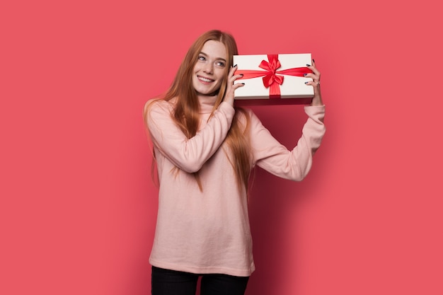 Rothaarige Frau mit Sommersprossen schüttelt ein Geschenk und lächelt auf einer roten Wand im Studio