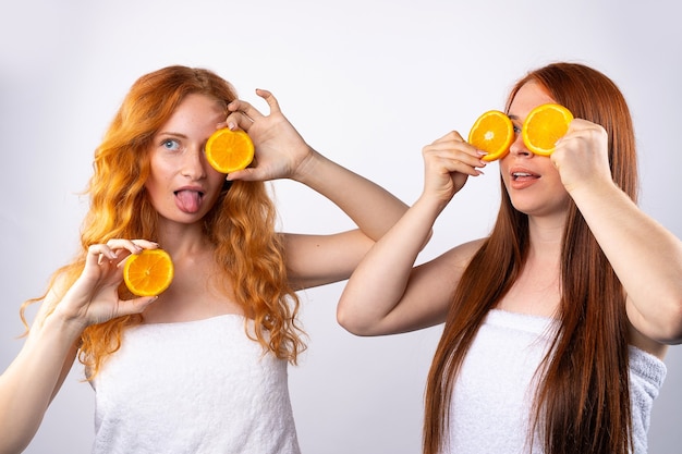 Rothaarige Freundinnen haben Spaß und posieren mit Orangenscheiben. Gesundes Essen, Spa und Schönheit, Vitamine und Glückskonzept. Foto auf weißer Wand.