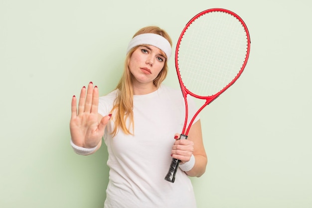 Rothaarige hübsche Frau, die ernsthaft aussieht und eine offene Handfläche zeigt, die ein Stoppgesten-Tenniskonzept macht