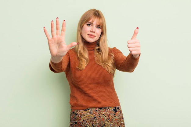 Rothaarige hübsche Frau, die lächelt und freundlich aussieht und Nummer sechs oder sechs zeigt, wobei die Hand nach unten zählt