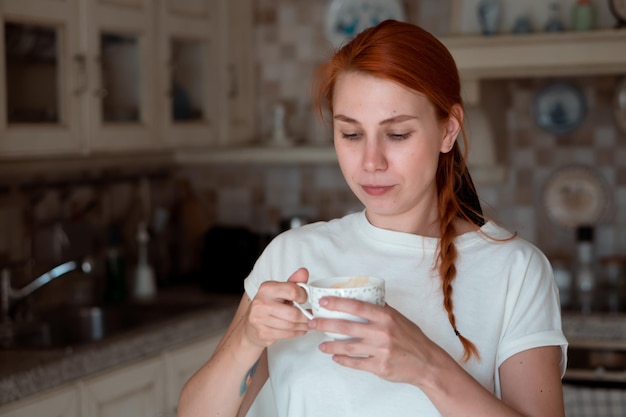 Rothaarige hübsche Frau, die zu Hause in der modernen Küche Kaffee trinkt