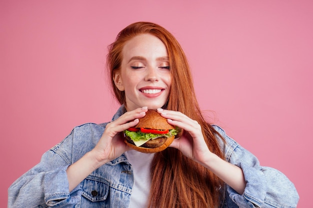 Rothaarige Ingwer mit Sommersprossen Frau genießt große riesige Burger Schnitzel und trägt demin amerikanische Jeansjacke im Studio rosa Hintergrund Traditionelles Konzept der USA