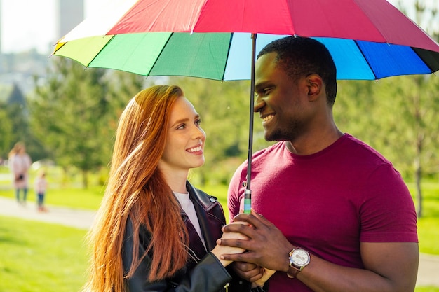 Rothaarige Ingwerfrau und afrikanisch-amerikanischer Mann stehen im Sommerpark nahe beieinander unter Regenschirm