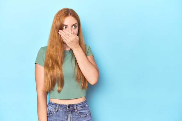 Rothaarige junge Frau auf blauem Hintergrund schaut nachdenklich auf einen Kopierraum und bedeckt den Mund mit der Hand