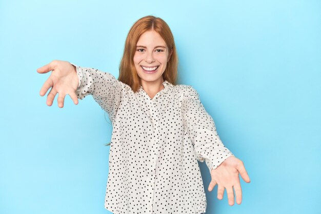 Rothaarige junge Frau auf blauem Hintergrund zeigt einen willkommenen Gesichtsausdruck