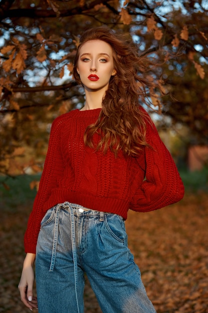 Rothaarige junge Frau in einem roten Pullover geht in den Park. Herbstschönheitsporträt einer modischen rothaarigen Frau bei Sonnenuntergang