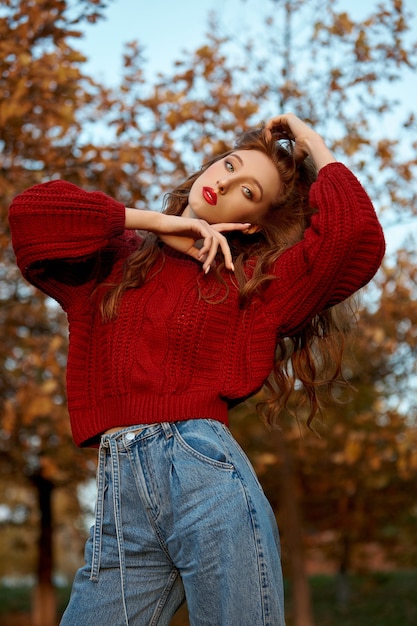 Rothaarige junge Frau in einem roten Pullover geht in den Park. Herbstschönheitsporträt einer modischen rothaarigen Frau bei Sonnenuntergang