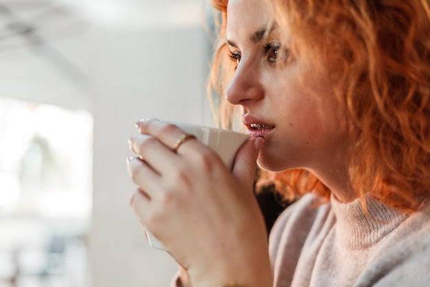 Rothaarige lockige junge helle Frau mit grünen Augen trinkt heißen Kaffee im gemütlichen Café Schönes Modell in stilvoller Kleidung Europa