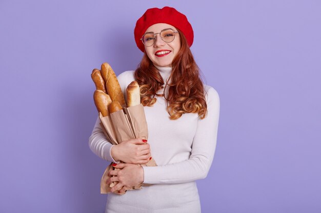 Rothaarige schöne lächelnde junge Frau, die Papiertüte mit Brot Baguette auf lila Raum hält