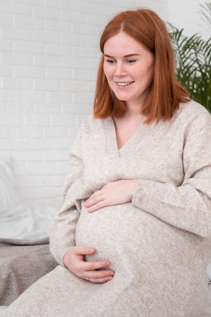 Rothaarige schwangere Frau in warmem, gemütlichem Kleid legt die Hände auf den Bauch und sitzt zu Hause im Schlafzimmer