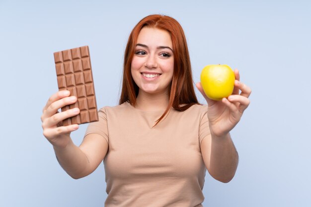 Rothaarige Teenagerfrau, die eine Schokoladentafel in einer Hand und einen Apfel in der anderen nimmt