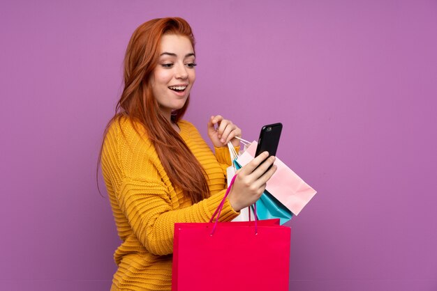 Rothaarige Teenagerfrau über isolierte lila Wand, die Einkaufstaschen hält und eine Nachricht mit ihrem Handy an einen Freund schreibt
