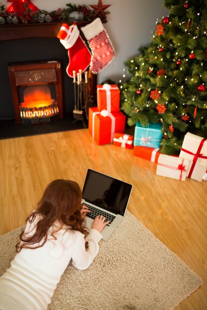 Rothaarigefrau, die auf Boden unter Verwendung des Laptops am Weihnachten liegt