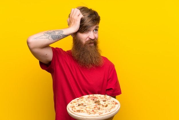Rothaarigemann mit dem langen Bart, der eine Pizza über der lokalisierten gelben Wand hat Zweifel und mit verwirren Gesichtsausdruck hält