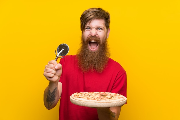 Rothaarigemann mit dem langen Bart, der eine Pizza über lokalisierter gelber Wand hält