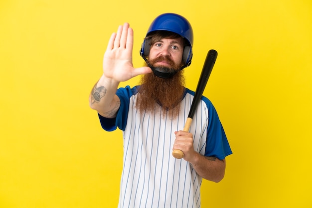 Rothaariger Baseballspieler Mann mit Helm und Schläger isoliert auf gelbem Hintergrund, der Stop-Geste macht