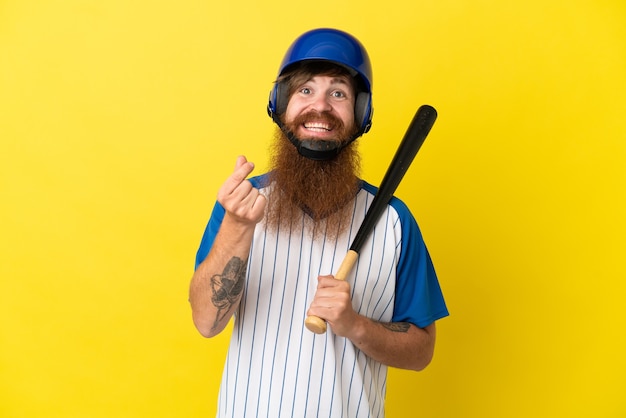 Rothaariger Baseballspieler mit Helm und Schläger isoliert auf gelbem Hintergrund, der Geldgeste macht