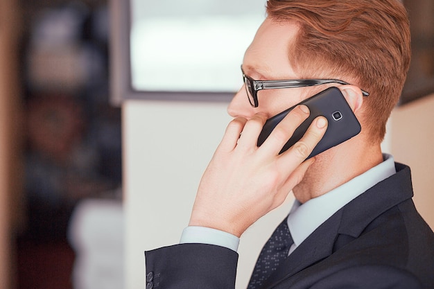 Rothaariger Geschäftsmann mit Brille spricht auf einer Handy-Nahaufnahme nach links. Kopf eines Mannes mit einem schwarzen Telefon am Ohr