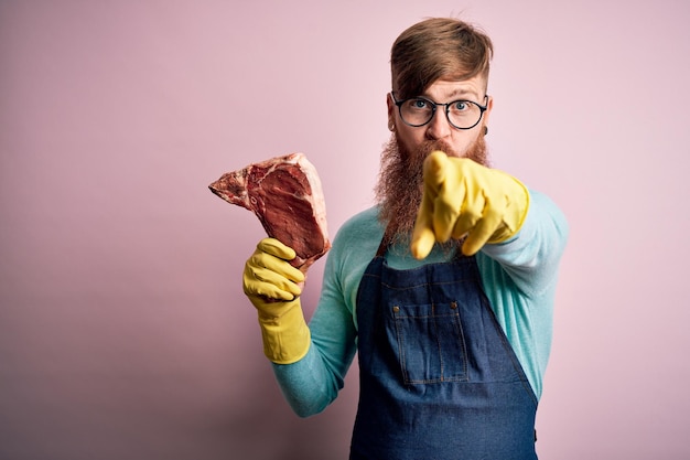 Rothaariger irischer Metzgermann mit Bart, der rohes Rindersteak über rosafarbenem Hintergrund hält und mit dem Finger auf die Kamera und auf Ihre Hand zeigt, positive und selbstbewusste Geste von vorne